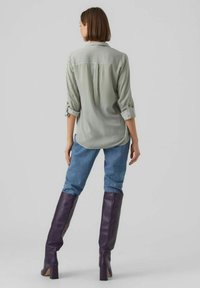 Gestreepte groene overhemd met opgerolde mouwen, gecombineerd met blauwe jeans en kniehoge paarse laarzen, gezien van achteren.