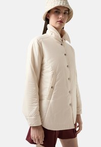 Beżowa kurtka pikowana z wysokim kołnierzem, zatrzaskami i bocznymi kieszeniami. Tkanina ma gładką teksturę i lekko błyszczące wykończenie.