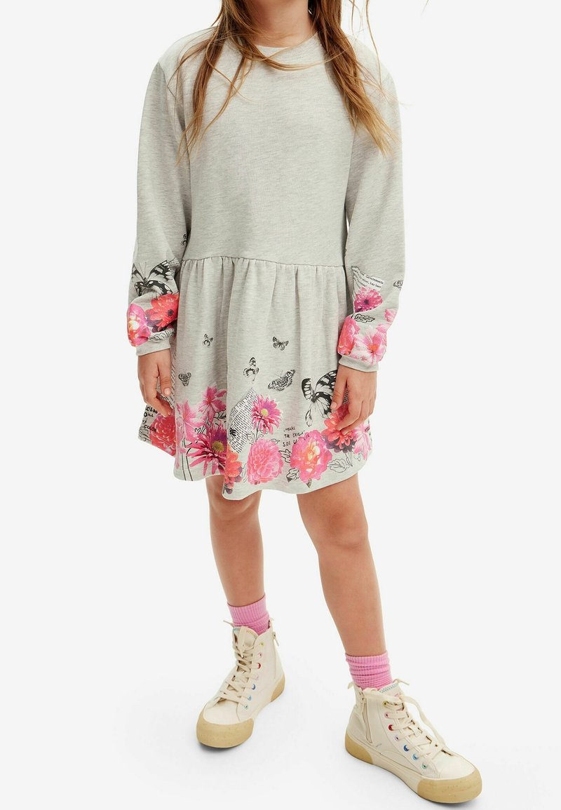 Jeune fille en robe gris clair avec imprimé floral et papillon rose vif, manches longues, baskets montantes crème et chaussettes roses.