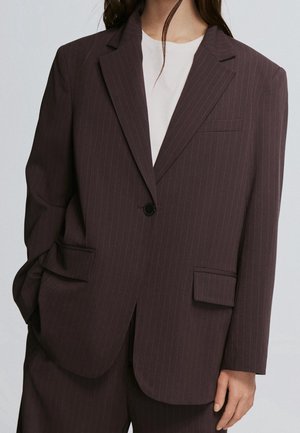 Kvinde iført en mørkebrun blazer med pinkstriber med en enkelt sort knap og matchende bukser, kombineret med en hvid top under.