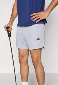Γκρι αθλητικό σορτς με υφή διχτυού και πλαϊνές σχισμές, με μαύρο λογότυπο Adidas. Συνδυασμένο με μπλε αθλητικό τοπ και μπαστούνι περπατήματος.