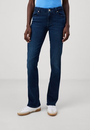 Personne portant un jean droit bleu foncé, des baskets blanches à semelles marron, et un haut bleu clair, debout sur un fond uni.