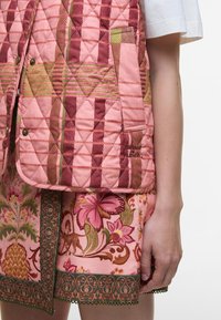 Colete pink acolchoado com padrões geométricos, combinado com uma saia estampada com flores que apresenta detalhes em verde e marrom e acabamento em rendinha decorativa.