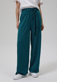 Pantalons larges turquoise avec une taille ceinturée, fabriqués à partir d'un tissu doux et fluide. Associés à des baskets blanches pour un look décontracté.