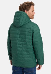 Mann mit kurzen Haaren, der eine grüne gesteppte Kapuzenjacke und blaue Jeans trägt, von hinten vor einem schlichten hellen Hintergrund gesehen.