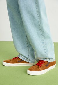 Zapatillas de ante marrón con cordones rojos y un acento marrón oscuro, combinadas con unos vaqueros claros de corte holgado sobre una superficie verde.