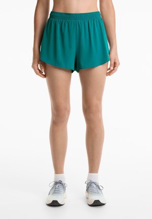 Türkise Sportshorts mit elastischem Bund, lockerer Passform und abgerundetem Saum. Kombiniert mit grauen Sneakers und weißen Knöchelsocken.
