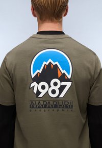 Olivově zelené tričko s velkým horským motivem v modré, černé a oranžové barvě, které zahrnuje rok "1987" a "NAPAPIJRI geographic" v černém písmu.