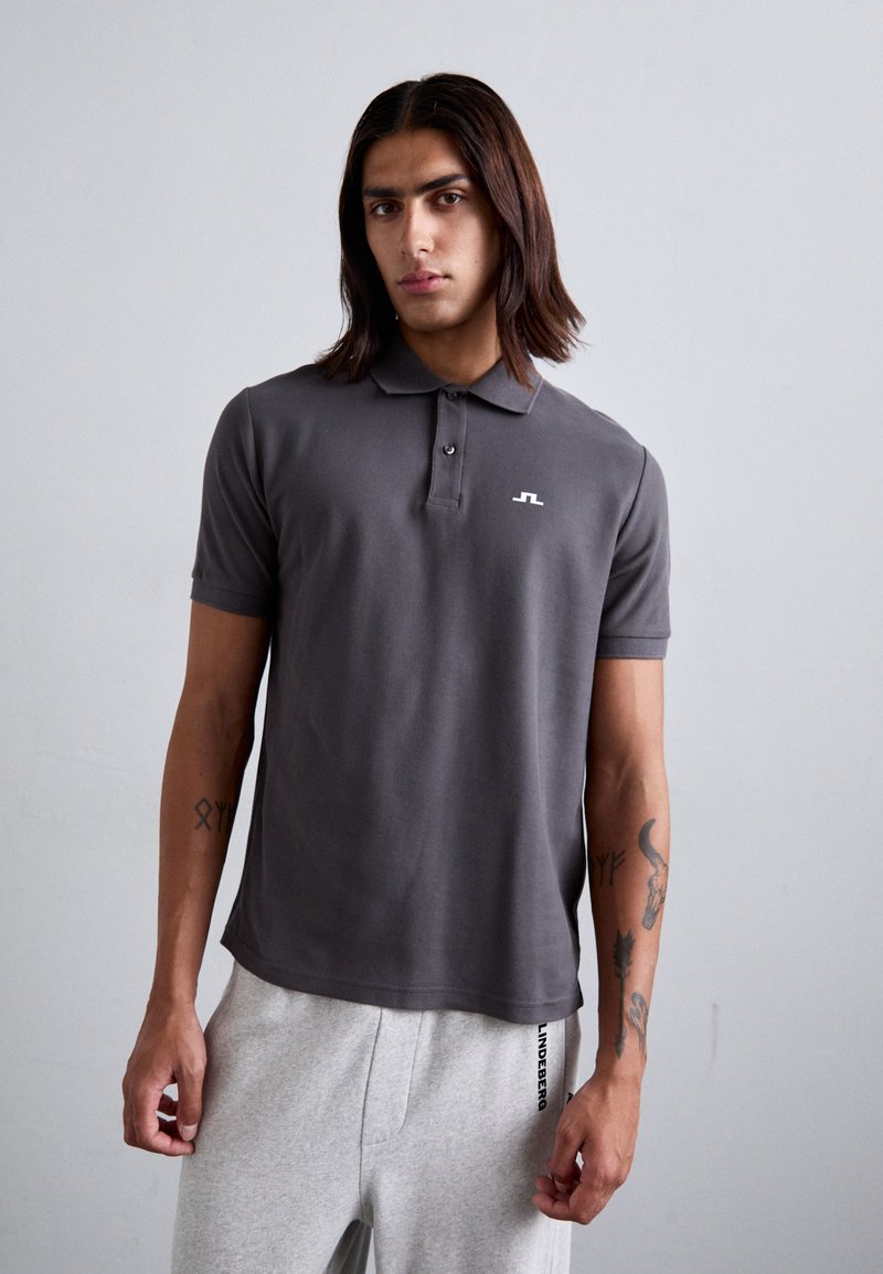 Graues Poloshirt mit kurzen Ärmeln, zwei vorderen Knöpfen und einem kleinen weißen Logo auf der linken Brust. Getragen mit grauen Jogginghosen, die seitlich beschriftet sind.
