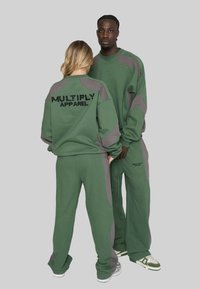 Zwei Modelle tragen grüne und graue Trainingsanzüge mit dem Schriftzug "Multiply Apparel", eines frontal und das andere zeigt den Rücken.