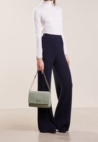 Borsa a tracolla in pelle verde con catena, forma rettangolare, texture liscia e dettaglio del logo in rilievo. Il modello indossa un dolcevita bianco e pantaloni a coste blu navy.