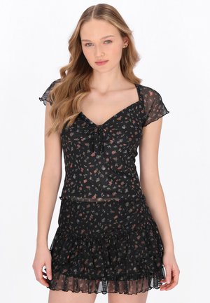 Ung kvinde med langt, bølget hår iført en sort blomsterprintet kortærmet kjole med flæsekant, stående foran en ensfarvet hvid baggrund.