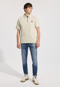 Homme debout portant un polo beige à manches courtes, un jean bleu retroussé et des baskets beiges sur un fond blanc uni.