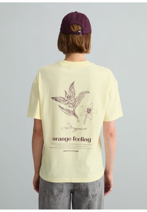 Person trägt ein hellgelbes T-Shirt mit braunen botanischen Skizzen und dem Text "orange feeling" auf dem Rücken, kombiniert mit grauen Jeans und lila Kappe.