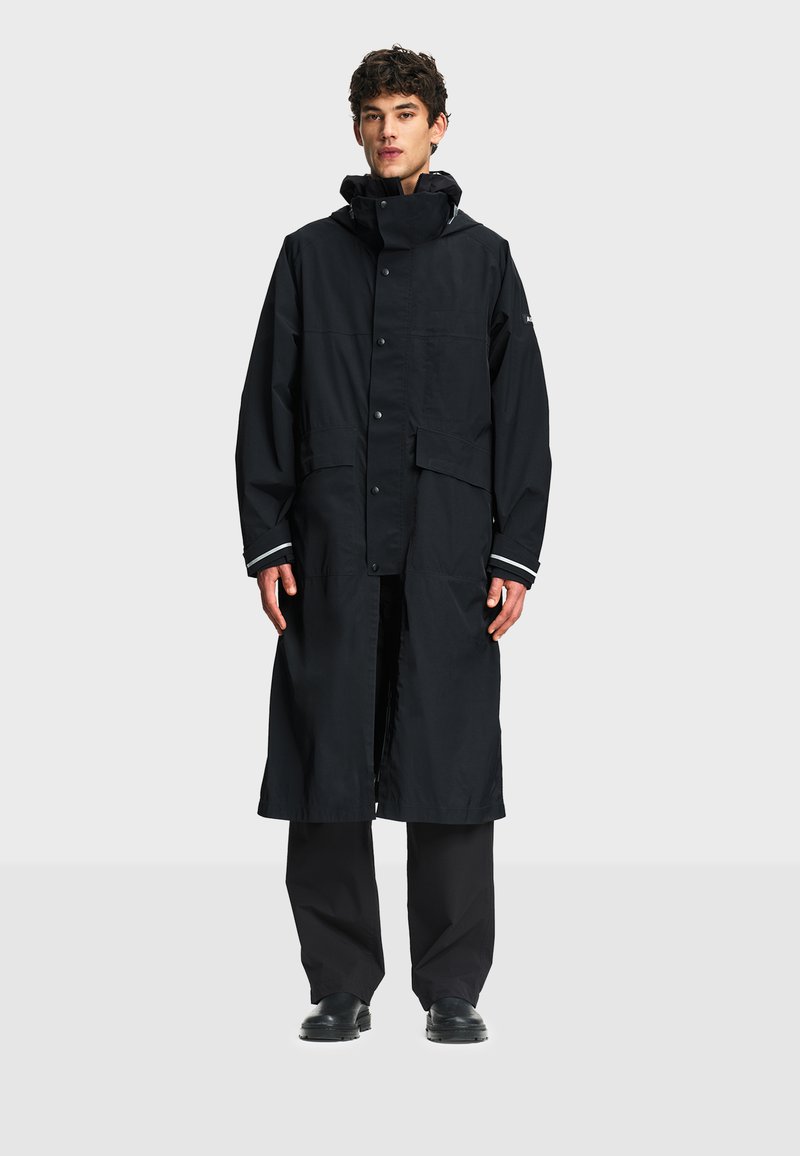 Homme debout portant un long imperméable noir avec fermeture à boutons, capuche, deux grandes poches avant, pantalon noir et chaussures noires.