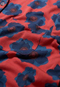 Czerwona tkanina z dużymi ciemnoniebieskimi wzorami kwiatowymi z wewnętrznymi detalami. Projekt zawiera kieszeń na piersi oraz kontrastowe przeszycia.