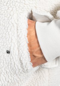 Mano parcialmente dentro de un bolsillo de una chaqueta de forro polar blanca y suave, con un botón a presión metálico visible y un puño de tela acanalada blanca.