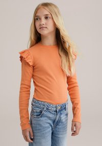 Szőke lány hosszú hajjal, narancssárga, puffos vállú, hosszú ujjú felsőt visel, világoskék farmernadrággal, és enyhén oldalra néz.
