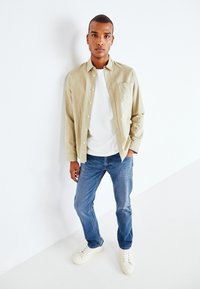 Lichtbeige button-up shirt, wit T-shirt, blauwe jeans en witte sneakers. Glad materiaal, ontspannen pasvorm, eenvoudig ontwerp met een zak.