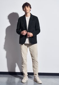 Černý kordurový blazer s klopami, v kombinaci se světle šedým tričkem a béžovými kalhotami. Model má na sobě béžové tenisky; pozadí z betonu.