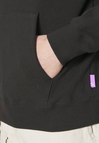 La main d'une personne repose dans la poche avant d'un sweat-shirt noir à manches longues avec une petite étiquette violette Puma visible.