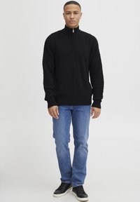 Mann steht frontal und trägt einen schwarzen strukturierten Strickpullover mit Halb-Reißverschluss, blaue Jeans und schwarze Sneaker mit weißen Sohlen.