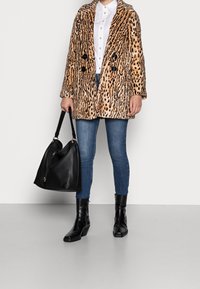 Persona vestida con abrigo de estampado de leopardo, camisa blanca de botones, jeans azules, botas negras hasta el tobillo, sosteniendo un gran bolso negro contra un fondo simple.