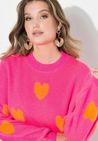 Jasnoróżowy sweter na drutach z dużymi pomarańczowymi wzorami w kształcie serca i prążkowanym, okrągłym dekoltem. Dopełnieniem są złote kolczyki stanowiące wyrazisty akcent.
