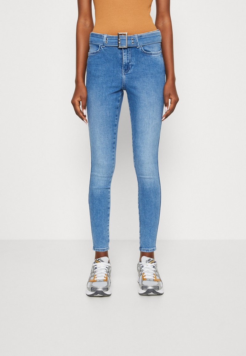Jeans de corte slim de color azul claro, con un cinturón a juego, que lleva una persona de pie, combinados con zapatillas blancas y grises.