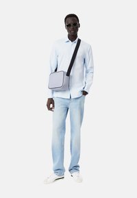 Bolso cruzado gris con correa negra, combinado con una camisa azul clara y jeans de lavado claro. El modelo lleva gafas de sol y zapatillas blancas.