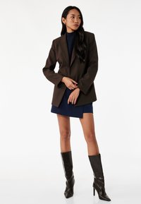 Bruine dubbelgeknoopte blazer van soepel materiaal, gedragen over een marineblauwe jurk. Model draagt zwarte puntige laarzen tot de knie, tegen een witte achtergrond.