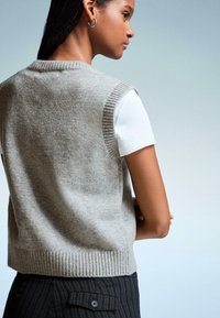 Graue gestrickte Weste mit gerippten Kanten, kurzen Ärmeln, über einem weißen T-Shirt, kombiniert mit schwarz gestreiften Hosen, von hinten gezeigt.