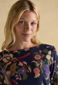 Koszula w kwiaty w kolorze granatowym z szerokim dekoltem; charakteryzuje się żywymi kwiatami, ptakami i liśćmi w odcieniach czerwieni, różu, złota i zieleni na fakturowanej tkaninie.