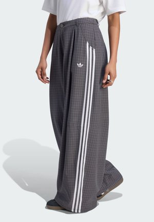 Persona que lleva pantalones de cuadros grises de pierna ancha con rayas verticales blancas y logo de Adidas, combinados con zapatos negros y camiseta blanca.