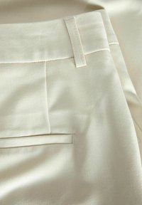Krämfärgad tyg med en slät yta, som har en platt midjeband, sidofickor och rena sömdetaljer.