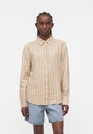 Μοντέλο με πλεξούδες, φοράει μπεζ ριγέ πουκάμισο με μακριά μανίκια και ανοιχτό μπλε τζιν σορτς, στέκεται μπροστά σε απλό φόντο.