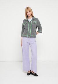 Cardigan corto grigio lavorato a maglia con dettagli verdi, due tasche frontali e scollo con colletto, abbinato a pantaloni larghi color viola chiaro.