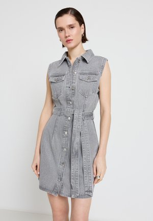 Junge Frau trägt ein ärmelloses graues Jeanskleid mit Knöpfen, Kragen und Taillengürtel, steht vor einem schlichten hellen Hintergrund.