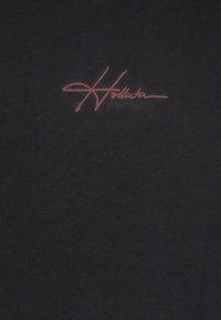 Černé tričko s jemnou texturou, které má výrazné červené nápisové logo "Hollister" na horní levé straně.