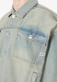 Svetlo modra jeans jakna z gumbnim zapiranjem, žepom na prsih, srebrnim okovjem in belim vezenjem na spodnjem delu.