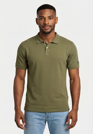 Uomo che indossa una polo verde oliva a maniche corte e jeans blu chiaro, in piedi di fronte a uno sfondo bianco.