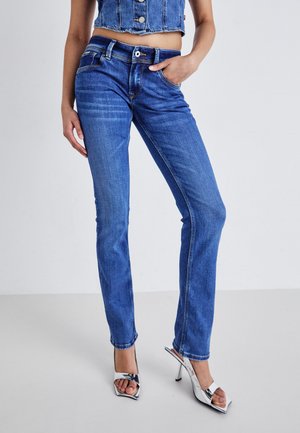 Jean en denim bleu avec une coupe droite, cinq poches et des coutures orange. Porté avec des sandales à talons hauts argentées.