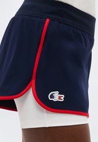 Laivastonsiniset urheilushortsit, joissa on punaiset reunukset tummalla iholla, valkoinen sisäkerros ja pieni krokotiililogo edessä.