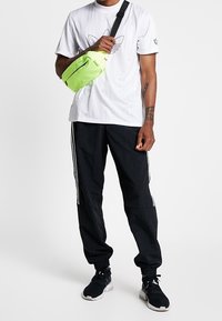 Grön lättviktskorsbodyväska med dragkedja, bärs över en vit t-shirt och svarta träningsbyxor med vita ränder samt svarta sneakers.