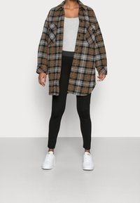 Koszula flanelowa w kratę w kolorze szarym, pomarańczowym i czarnym z dwiema kieszeniami na piersi, noszona na szarym topie i czarnych legginsach, zestawiona z białymi sneakersami.