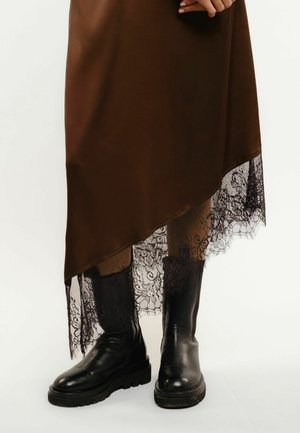 Brauner asymmetrischer Rock mit schwarzem Spitzenbesatz, kombiniert mit schwarzen Lederstiefeln bis zur Mitte der Wade, auf hellem Hintergrund.