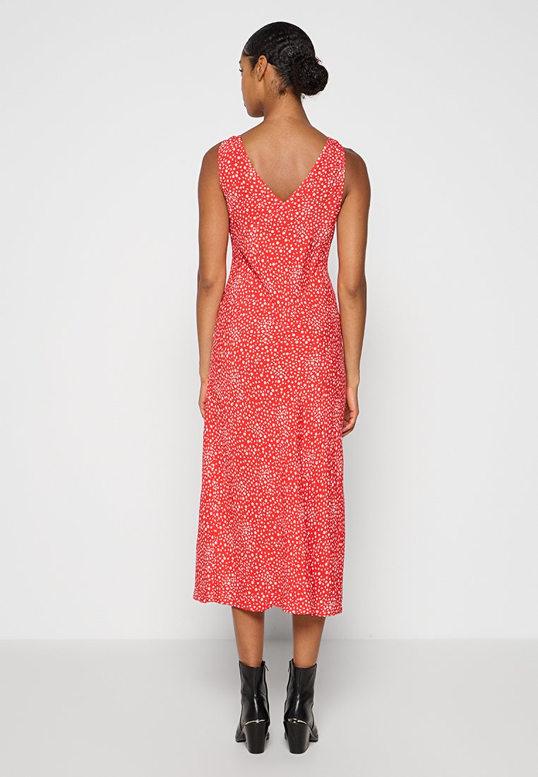 Abito rosso di lunghezza media con pattern a pois bianchi, senza maniche, con scollatura a V sul retro e tessuto fluido. Indossato con stivali neri alla caviglia.