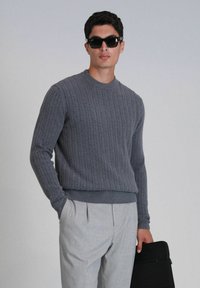 Grauer Kabelstrickpullover mit Rundhalsausschnitt, kombiniert mit hellgrauen, plissierten Hosen; hält ein schwarzes Portfolio. Glatte Textur.