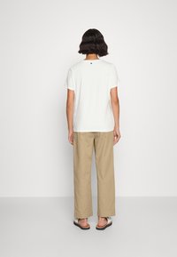 Witte T-shirt met korte mouwen en een klein zwart logo op de achterkant, gecombineerd met losse beige broek en open teen sandalen.