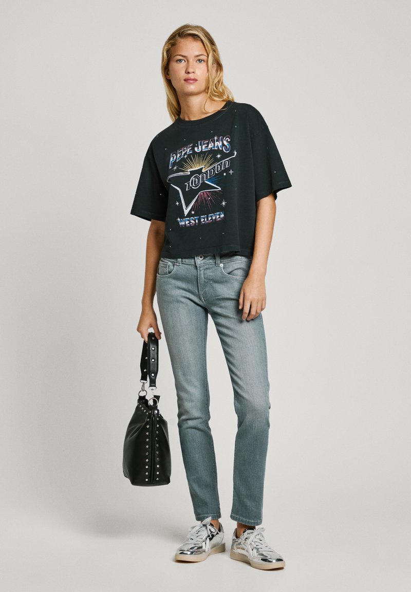 Maglietta nera crop con grafica colorata e testo, abbinata a jeans azzurri chiari. Accessoriata con una borsa nera e scarpe da ginnastica argentate.